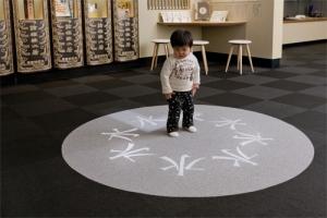 「大漢和辞典」の書棚の前に円形に「水」を書かれたカーペットに立つ幼児の写真