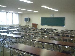 長机と折り畳み椅子が並び、黒板とテレビモニターのある講義室の写真
