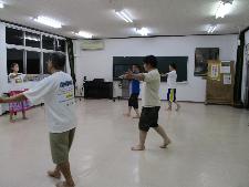 室内で女性講師1人と男性4人がフラダンスの練習をしている。